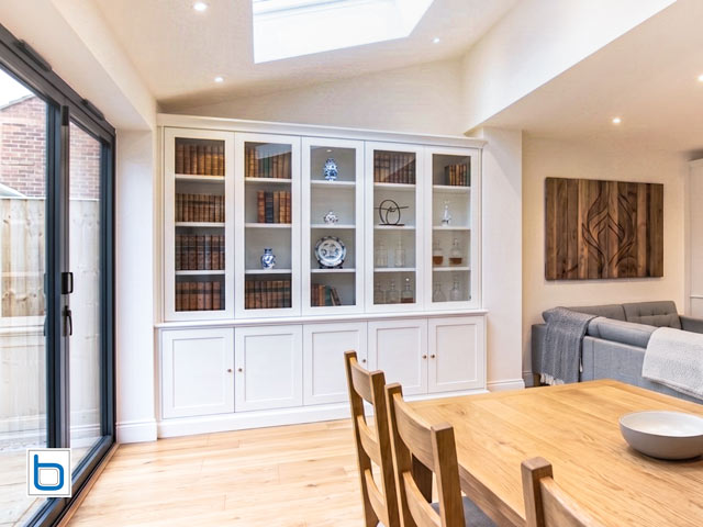 A large custom built-in white dining room dresser featuring five shaker base cupboards beneath tall glass-fronted display cabinets filled with books and ceramics in a modern home extension.