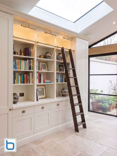 A grand custom built-in white dining room bookcase featuring shaker base cupboards and drawers supporting multi-tier open shelves, complete with brass picture lights and a dark wood rolling library ladder.
