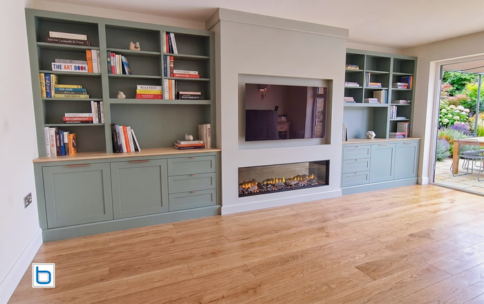 Fitted living room furniture in a sage green (F&B Suffield Green + ammonite chimney breast) false chimney breast with electric fire, TV recess, and shaker bookcases with oak worktop