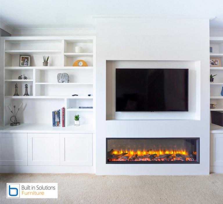 Bespoke White living room furniture featuring lower shaker built-in cupboards, open display shelves, a recessed television, and an integrated modern electric fireplace.