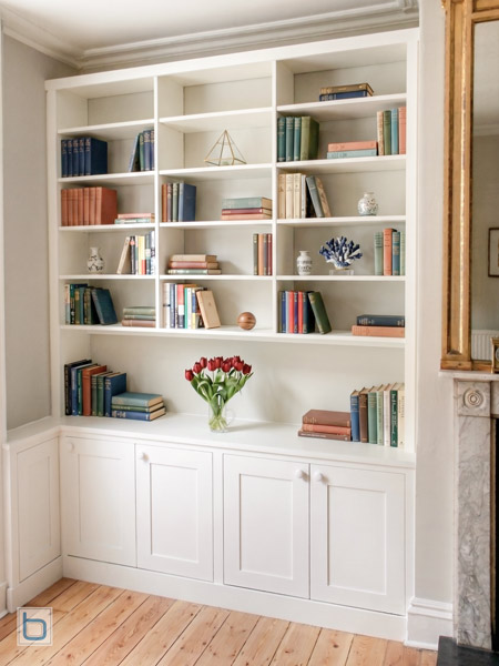 Bespoke white built-in cabinets showcasing a custom corner wrap layout, traditional lower shaker storage cupboards, and a wide open display library shelving system besides a marble fireplace.