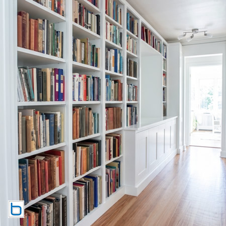 Bespoke white built-in cabinets configured as a long hallway library wall featuring floor-to-ceiling open bookshelves and low-level shaker storage cupboards.
