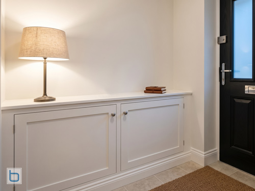Custom white built-in cabinets configured as a low-level hallway side cupboard featuring shaker doors, metal knobs, and a table lamp on top next to a front door.