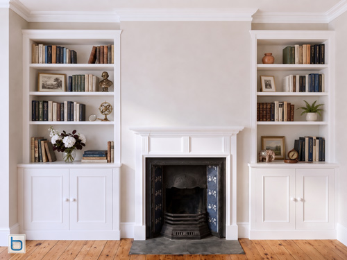Bespoke white built-in cabinets featuring shaker door alcove cupboards and open display bookshelves positioned besides a traditional period fireplace.