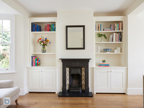 Bespoke white built-in cabinets featuring symmetrical shaker door alcove cupboards and open display bookshelves positioned besides a traditional period fireplace.