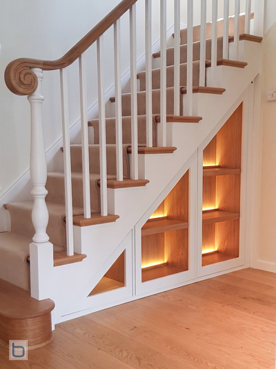 Bespoke white fronted built-in cabinets configured as under-stairs open display shelves featuring a warm oak interior, wooden shelving, and integrated LED backlighting.