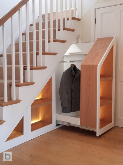 Bespoke white Slide out built-in Bookcase cabinets showing an under-stairs pullout storage unit extended out from the framework to reveal integrated oak shelves and warm LED lighting.