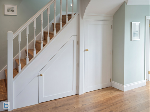 Bespoke white built-in cabinets configured for under-stairs storage featuring custom angled shaker doors and brass knobs.