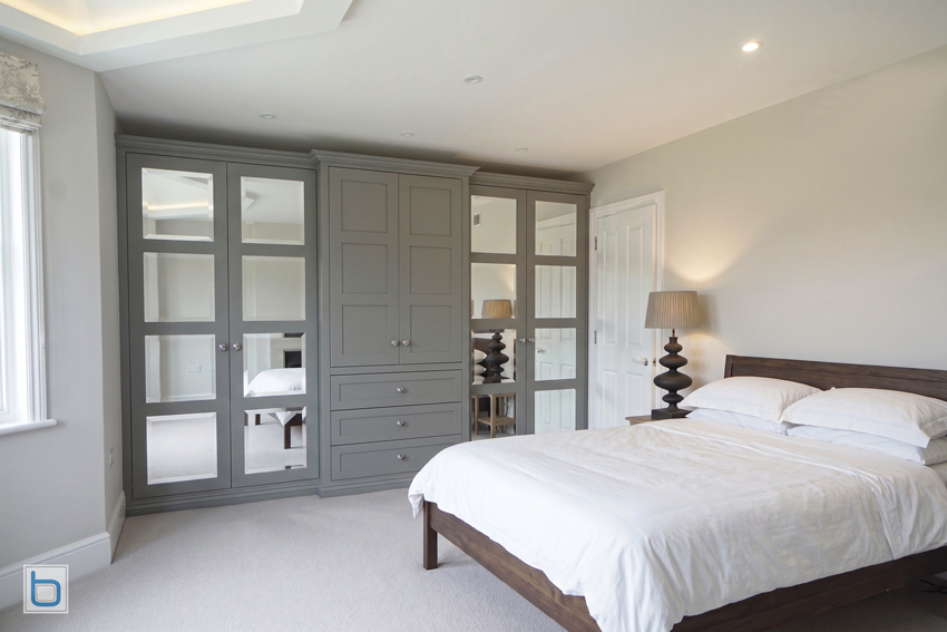 Bespoke grey Victorian wardrobes featuring four-panelled doors with premium bevelled edge mirrors, flanking a prominent central chest of drawers and cabinet next to a dark wood bed frame.