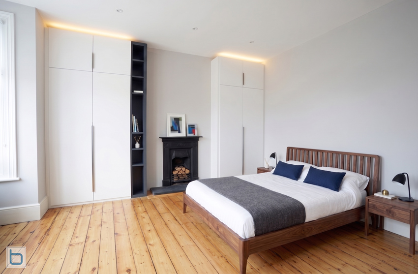 Symmetrical White contemporary wardrobes featuring long vertical handles, matching top cupboards, and integrated warm overhead lighting framing a central fireplace with built-in dark Hague Blue shelving.