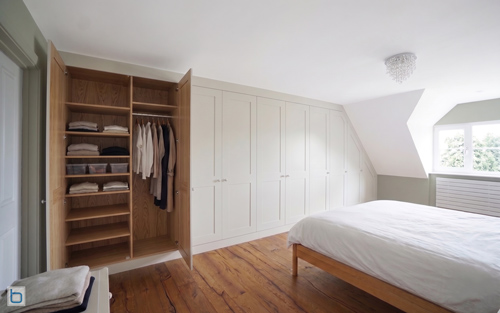 A premium fitted wardrobe installation spanning a sloped bedroom wall, showing open doors that reveal a warm oak timber carcass interior with custom shelves, clothing rails, and organized garments.