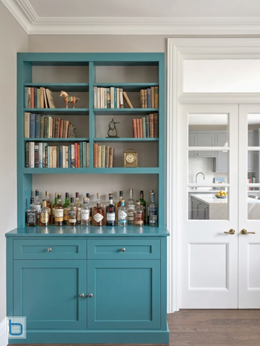 A custom built-in teal home bar and dresser unit featuring a two-door shaker base cabinet with drawers supporting an open counter filled with liquor bottles beneath multi-tier bookshelves.