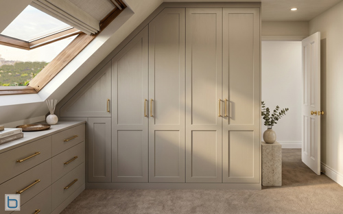 Custom-built loft wardrobes precisely fitted under an angled ceiling, featuring classic warm beige Shaker doors, elegant gold bar handles, and a matching chest of drawers beneath a skylight.