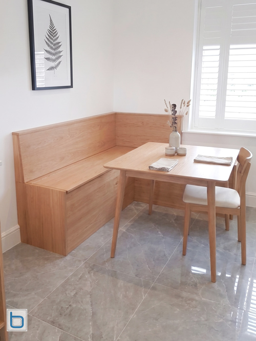 A custom-built L-shaped natural oak corner banquette seating bench with a high wooden backrest, positioned next to a matching oak dining table and chairs on a grey tiled floor.