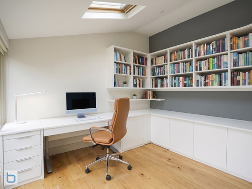 A modern custom built-in L-shaped home office desk and bookcase unit in white, featuring handleless base storage cupboards, a drawer line unit, and floating corner bookshelves under a sloped ceiling with a roof window.