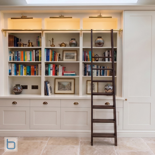 A premium white home library featuring a dark wooden ladder on a polished brass rail, warm integrated LED shelf lighting, overhead brass picture lights, and a shaker cupboard base.