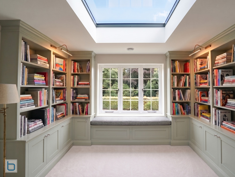 A custom floor-to-ceiling sage green home library flanking a central window seat under a large skylight, featuring detailed cornicing and inset panel base cupboards.