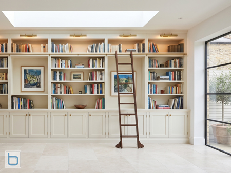 Cream painted floor-to-ceiling fitted home library with brass picture lights, wooden library ladder on rail and glazed base cupboards