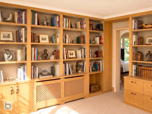 A luxury custom-built solid oak home library wrapping around a room, featuring floor-to-ceiling bookshelves with Farrow & Ball Ammonite painted backing panels, integrated diamond-lattice radiator covers, and built-in lower cabinetry.
