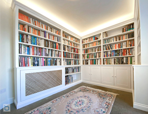 A custom white wrap-around corner home library featuring floor-to-ceiling bookshelves, a built-in diamond-lattice radiator cover, traditional shaker base cabinets, and warm uplighting from the cornicing.