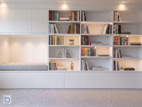 A modern custom-built white home library wall with handleless push-to-open base cabinets, asymmetric flat open shelving with integrated LED backlighting, and a cosy built-in reading daybed nook.