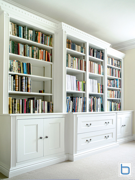 A grand white bespoke breakfront home library with dentil cornicing, fluted pilasters, adjustable open upper bookshelves, and traditional shaker base cabinets with drawers.