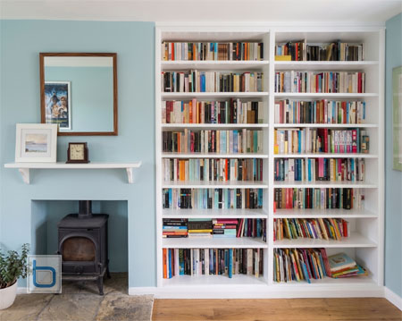 Bespoke floor-to-ceiling white painted open bookcase fitted flush into a living room recess next to a wood-burning stove.