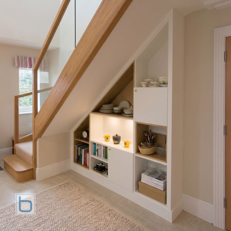 Modern custom white under-stairs cupboards and open display shelves with ambient LED lighting under an oak and glass staircase.