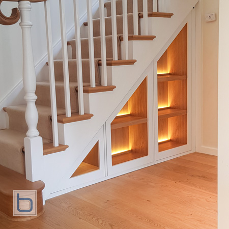 Bespoke white under-stairs storage unit with warm integrated LED strip lighting and natural oak backing.