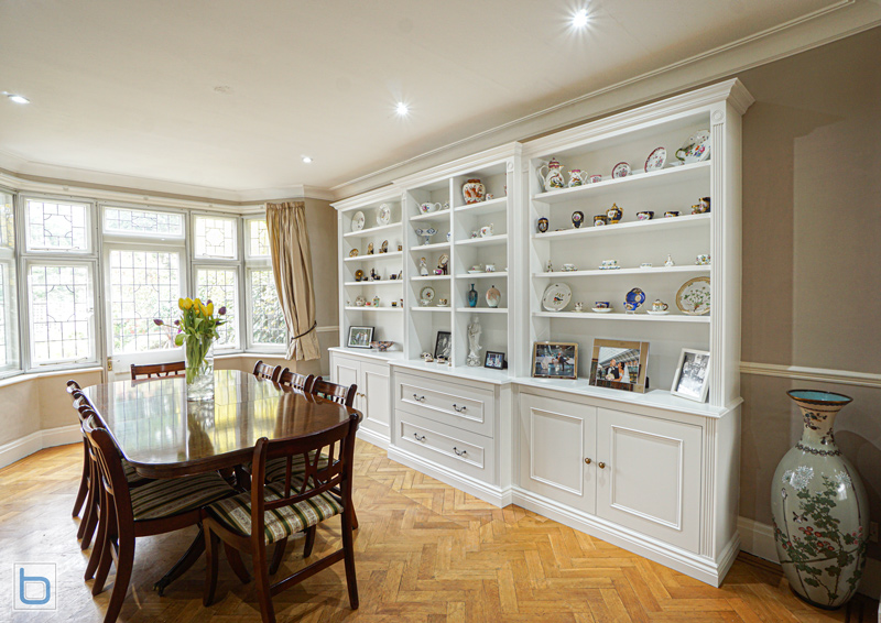 A large custom built-in white dining room display unit featuring shaker base cupboards, wide central drawers, and multi-tier open shelves showcasing fine china, set against a classic herringbone parquet floor.