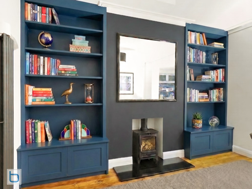 Bespoke built-in alcove bookcases painted in Farrow and Ball Hague Blue with lower cupboards and open shelving, besides a central chimney breast painted in dark grey featuring a black wood burning stove.