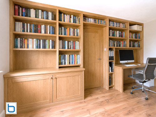 A grand custom built-in natural oak library and home office unit spanning an entire wall, featuring shaker base cupboards, a fitted desk station, and multi-tier open shelves precision-engineered to encase a central matching timber doorway.