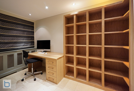 A bespoke natural wood study library featuring a floor-to-ceiling open cubby bookcase integrated alongside a matching timber desk with drawers, situated in a modern home office with stone flooring.