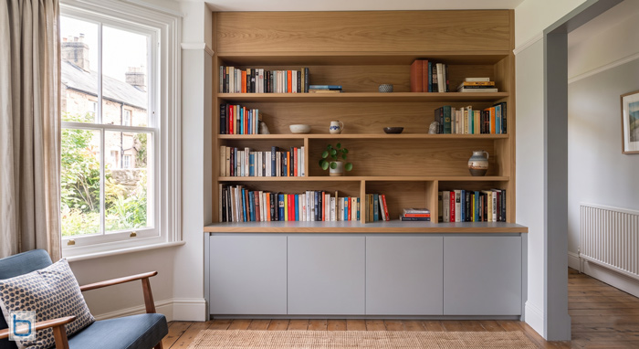 A sample photo showcasing a custom built-in contemporary wall unit with open oak bookshelves above a grey handleless storage sideboard, positioned next to a traditional sash window.