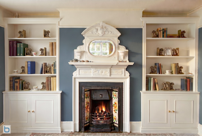 A sample photo showcasing custom built-in alcove shelving units with lower Shaker style storage cupboards on either side of an ornate fireplace featuring an integrated overmantel mirror.