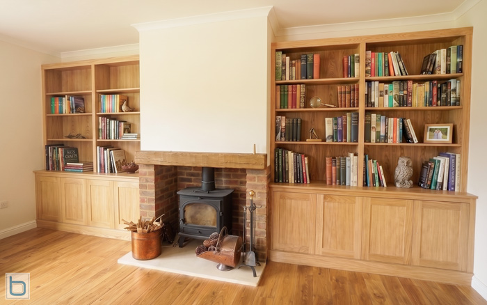 A sample photo showcasing custom built-in alcove shelving units made from natural grain oak timber, featuring open upper bookshelves and lower storage cupboards on either side of a brick fireplace with a wood-burning stove.