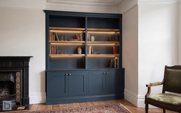 A sample photo showcasing a custom built-in living room bookcase unit painted in a dark blue hue, featuring illuminated oak shelves above classic Shaker style lower cupboards.