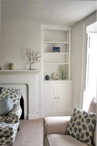 A custom-built white alcove unit featuring a traditional two-door shaker base cupboard with small knob handles beneath open display shelves supporting books and ornaments.