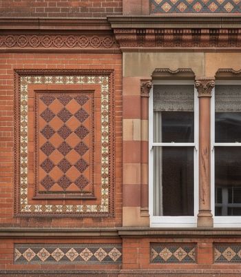 A close-up photograph of detailed Victorian brickwork on a building facade in Hungerford. The exterior architecture shows red bricks alongside intricate terracotta diamond tiles and decorative stone borders. On the right, a traditional white wooden sash window is framed by two carved stone columns with detailed capitals, reflecting the historic building and masonry styles of the Victorian era.