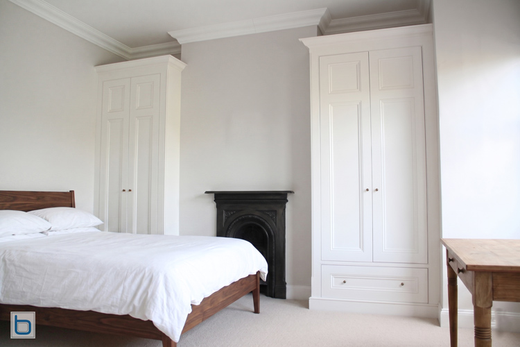 Alt Text: A pair of white built in Victorian wardrobes flanking a black cast iron fireplace, one with panelled doors and a base drawer, in a bedroom with a wooden bed frame.