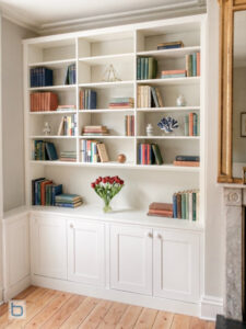 Bespoke white built-in cabinets showcasing a custom corner wrap layout, traditional lower shaker storage cupboards, and a wide open display library shelving system besides a marble fireplace.