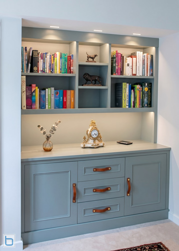 A custom-built alcove dresser unit painted in Farrow & Ball Pigeon green-grey, featuring inset shaker base cupboards with leather pull handles, a central stack of drawers, and open upper display shelving.