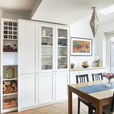 A bespoke white fitted dresser in a dining room with glass fronted upper cabinets, closed base cupboards, and a built in wine rack beside a wooden dining table.