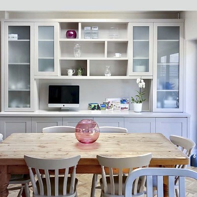 A bespoke fitted dresser unit in a farmhouse kitchen, painted pale grey with glass fronted display cabinets above and shaker cupboards below a farmhouse table.