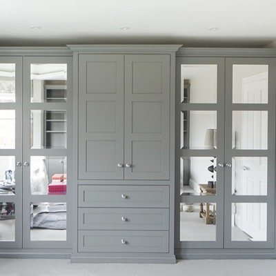 Bespoke fitted wardrobes in a soft grey finish with mirrored door panels either side and a central bank of drawers with chrome handles in a modern bedroom.