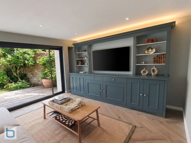A sample photo showcasing a custom built-in media wall painted in a dark blue-grey hue, featuring lower storage cupboards, drawers, open display shelves, and a large flat-screen television next to sliding glass doors.
