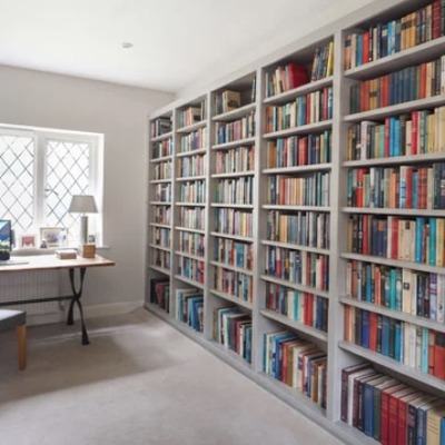 Bespoke fitted oak bookcases lining the walls of a home office, filled with books, beside a small writing desk near a window.