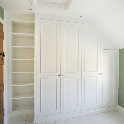 Bespoke white fitted wardrobes built into a sloped loft ceiling, combining shaker doors with open shelving to one side for extra storage
