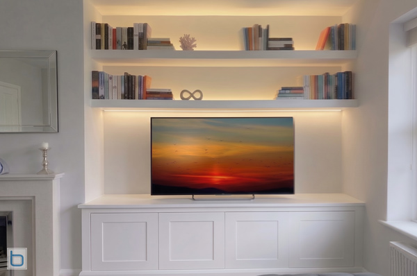 A custom-built white media wall unit fitted into a wide alcove, featuring a four-door shaker base cabinet supporting a flat-screen television beneath thick floating open shelves with warm integrated LED accent lighting.