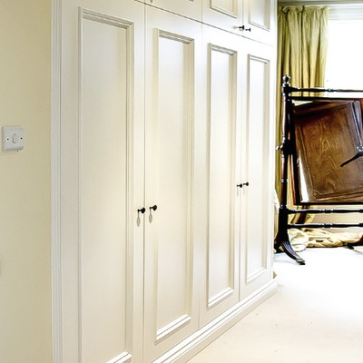Close-up of cream fitted wardrobe doors with black knob handles, positioned beside a window in a traditional bedroom.
